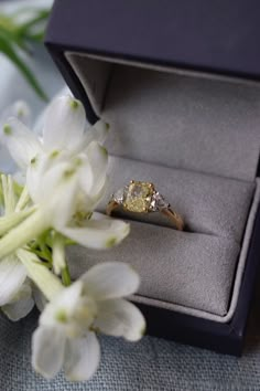 a yellow diamond ring sits in an open box next to white flowers on a table