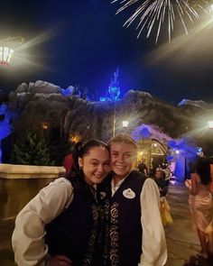 two people standing in front of a castle at night with fireworks above them and onlookers