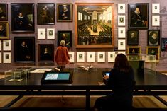 a woman sitting at a table in front of a wall full of paintings and books