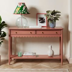 a pink console table with two vases and a lamp on the top, in front of a white wall