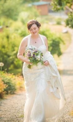 a woman in a wedding dress walking down a dirt road holding a bouquet and looking at the camera