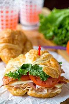 a sandwich with meat, lettuce and tomato sitting on wax paper next to some croissants