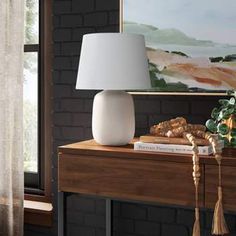 a white lamp sitting on top of a wooden table next to a painting and window