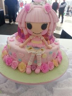 a pink cake with a doll on top and flowers around the edges, sitting on a table