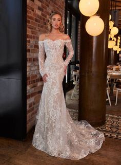 a woman standing in front of a brick wall wearing an off the shoulder wedding dress