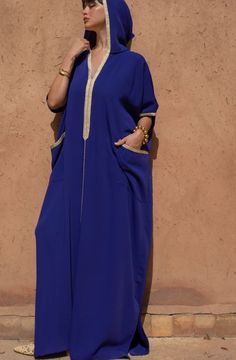 Experience the epitome of Moroccan fashion with our Oversized Crêpe Hooded Jalabiya in a soothing Blue hue. This jalabiya seamlessly combines tradition and modernity, creating a statement piece that's both comfortable and stylish.   Product Details:      Oversized Jalabiya : This jalabiya boasts an oversized fit, providing roominess and comfort for all body types.     Golden "Sfeefa" Details : Adorned with golden "sfeefa" detailing on the neckline, sleeves, and spacious front pockets, this jalab Halston Kaftan, Blue Tunic Thobe For Eid, Jalabia Styles For Women, Hooded Kaftan, Somali Clothes, Moroccan Dresses, Moroccan Kaftan Dress, Morocco Fashion, Moroccan Clothing