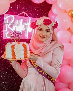 a woman in hijab holding up a cake with candles on it and balloons behind her