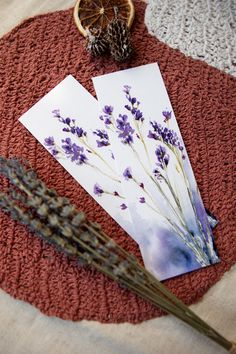 two cards with dried lavenders and an orange slice on a red mat next to them