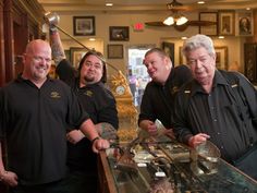 five men in black shirts are standing behind a counter with silverware on it and one man is pointing at the camera