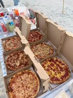 four pizzas in boxes sitting on a table at an outdoor picnic area with drinks and condiments