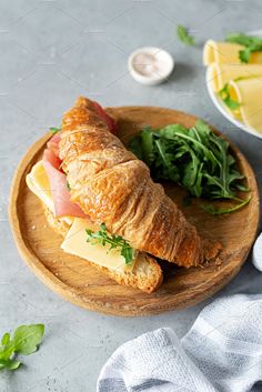 a croissant sandwich with ham and cheese on a wooden plate next to fresh greens