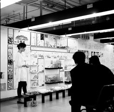 two people standing in front of a wall with pictures on it and one person sitting at a table