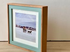 two people and a dog are standing in the snow with mountains behind them on a wooden frame