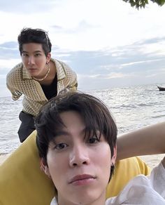 two people sitting on the beach with one person leaning over his head and another standing behind him