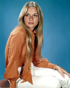 a woman sitting on top of a wooden table next to a blue wall in an orange shirt and white pants