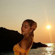 a woman standing in front of the ocean with her eyes closed and hands behind her head