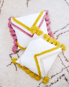 two white pillows with yellow and pink tassels laying on top of a rug