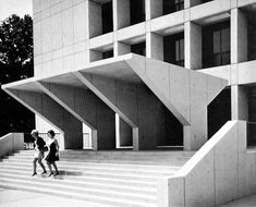 black and white photograph of two people walking up stairs in front of a building with large windows