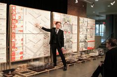 a man in a suit standing next to a wall with plans on it and people looking at them