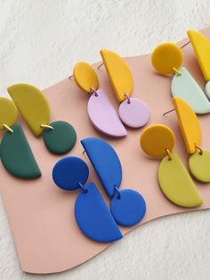 several pairs of colorful earrings on a pink surface with white paper behind them and the bottom half of each earring has been cut out