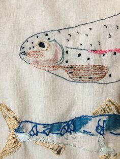 two embroidered fish on white fabric with blue and pink threads in the bottom right corner