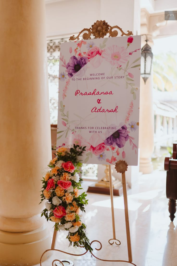 a sign with flowers on it sitting in front of a pillar at a wedding ceremony