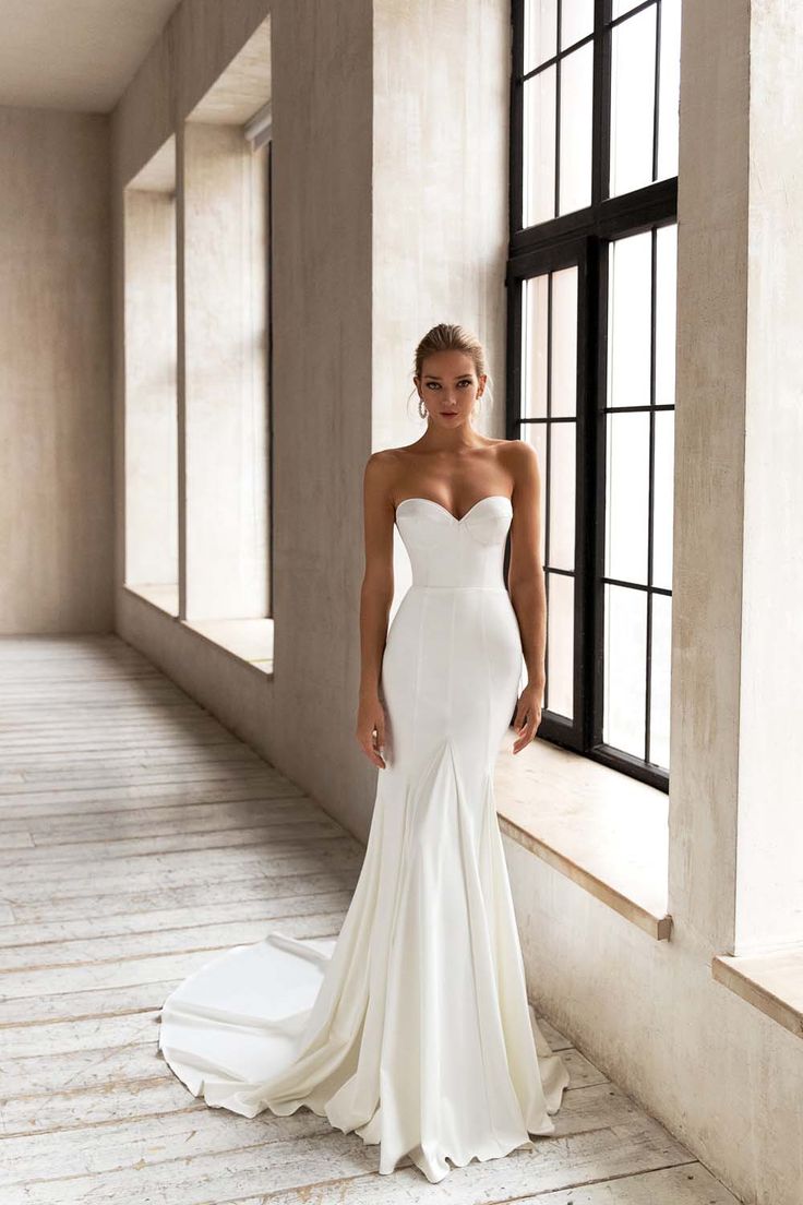 a woman in a white wedding dress standing next to a window and looking at the camera