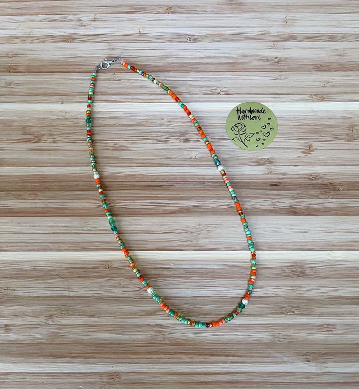 an orange and green beaded necklace sitting on top of a wooden table next to a tag