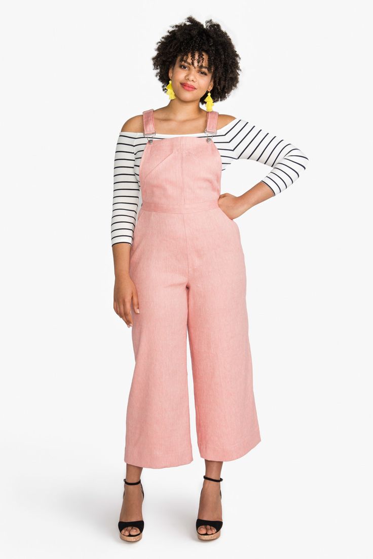 a woman standing in front of a white background wearing pink overalls and striped shirt