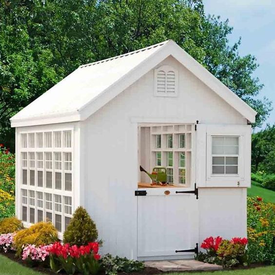 a small white shed sitting in the middle of a garden with lots of flowers around it