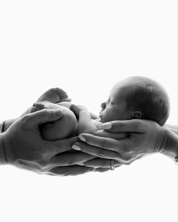 black and white photograph of a baby being held by two adult's hands in front of a window