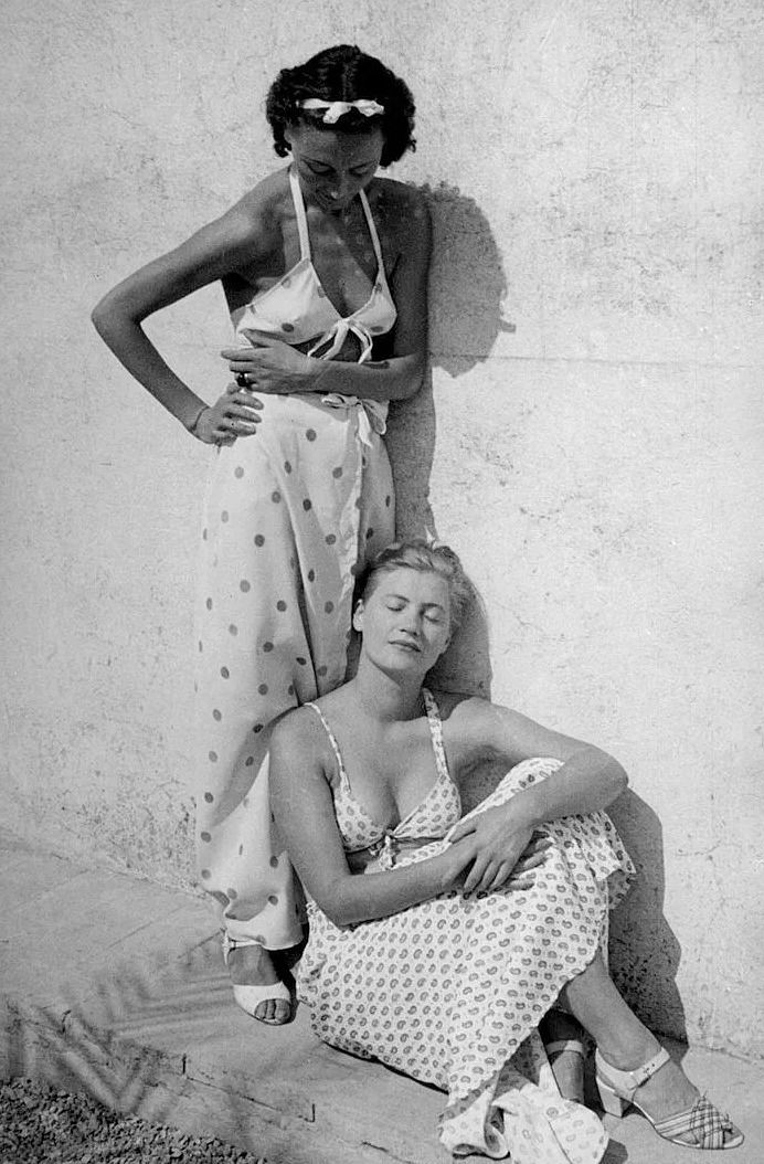 two women in bathing suits standing next to each other on the ground with their arms around one another