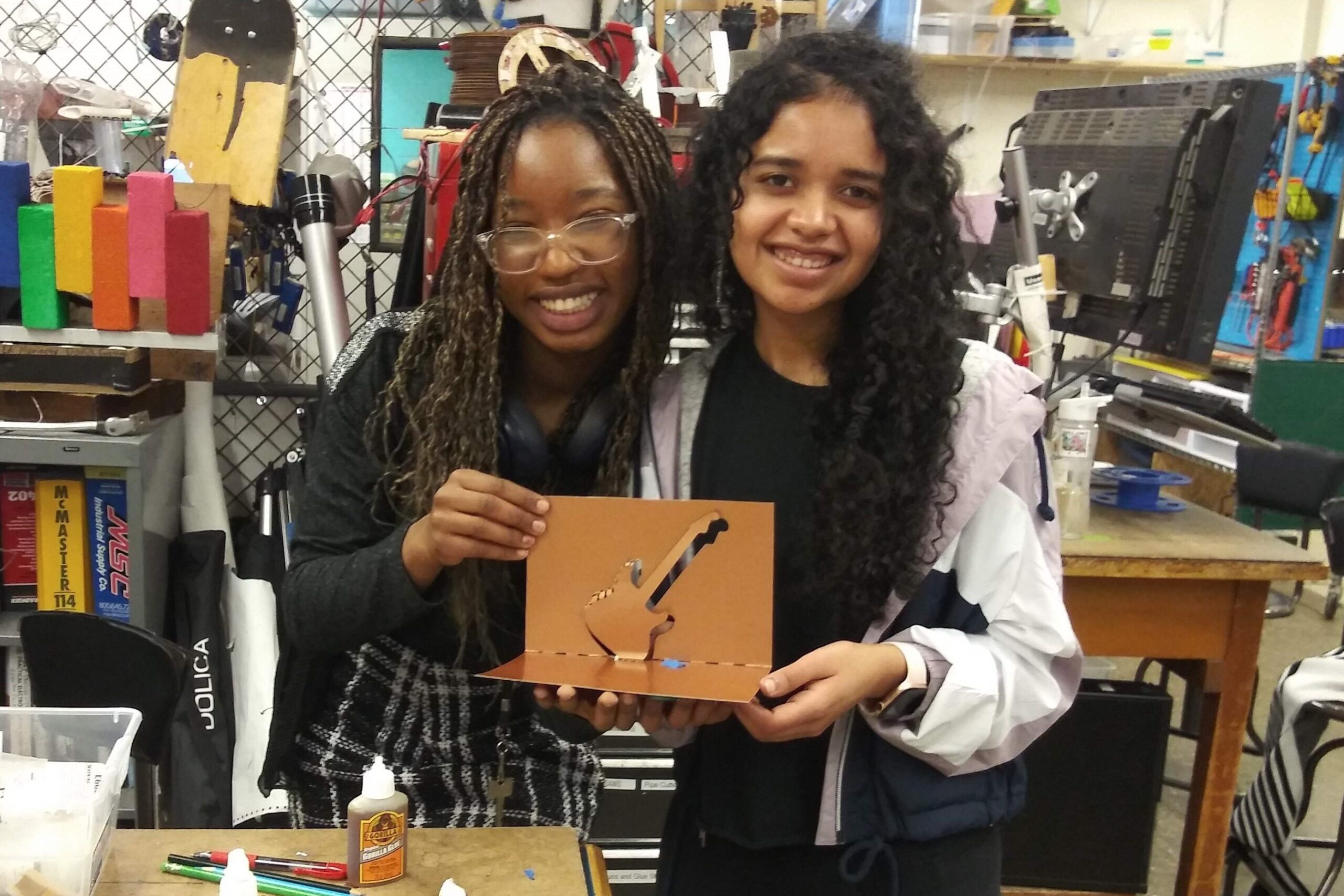 Two people proudly holding up a paper-cut project. They are surrounded by classroom tools.