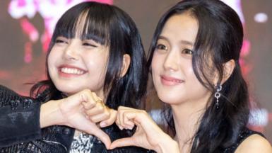 Two women, looking to the left of the camera, join their hands to make a heart shape and smile at a red carpet event