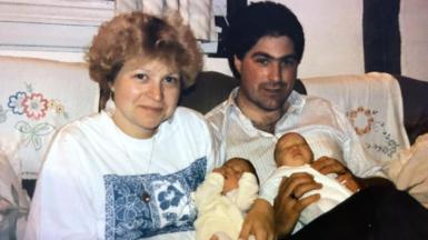 Angela Roberts and her husband Martin Roberts sat on a sofa holding their twin daughters Nikki and Becky in 1991. Angela has blonde permed hair, long earrings and a necklace, and is wearing a white T-shirt with blue floral motif. Martin has black hair and is wearing a white striped shirt.