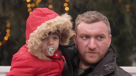 A man holds a young child who is wearing a bright red coat with hood, in the snow