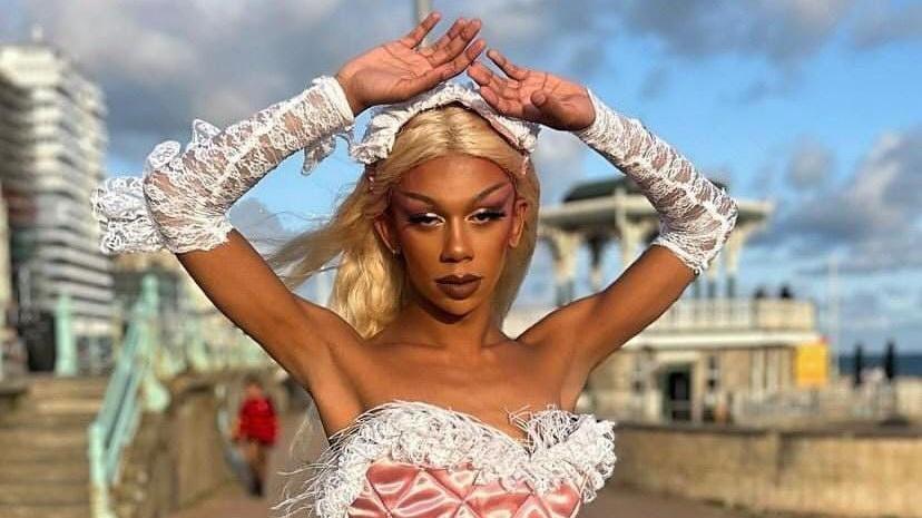 Kane, as Raiine, pictured outside on Brighton sea front. Raiine wears a pale pink bustier top with a white ostrich feather trim and matching white lace gloves. She wears a long blonde wig and raises her arms above her head. 