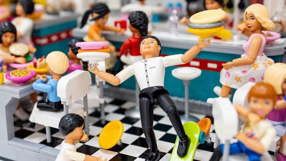 A busy 1950s diner scene made from Lego showing a waiter slipping on a skateboard