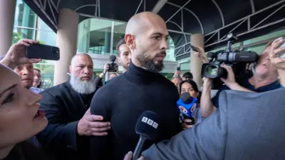 Andrew Tate, bearded and wearing a black sweater, is surrounded by press outside of an airport terminal