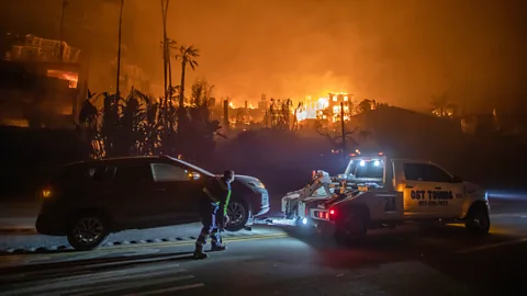 An abandoned car is hitched to a tow truck while buildings burn in the background (Credit: Getty Images)