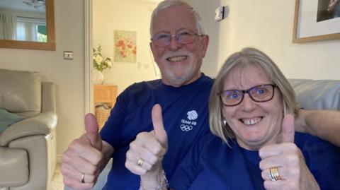 Meg Richards sits alongside husband Peter, sticking their thumbs up and smiling as they wear purple team GB shirts