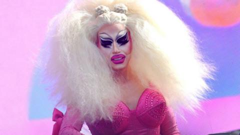 Trixie Mattel performing a DJ set. Trixie wears an enormous blonde wig and exaggerated make-up, including bright pink lips and black eyeliner. She wears an embellished bright pink corseted dress 