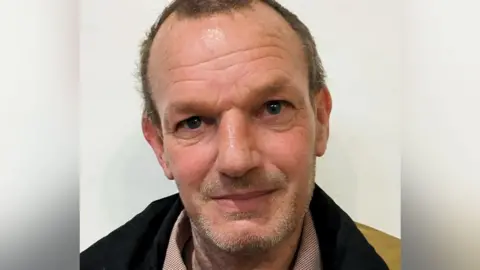 A close-up of a Nick Starmer, who has light skin and short hair. His expression is neutral but slightly warm, with a faint smile. The background is plain and white. He is wearing a dark jacket and a light-coloured shirt.