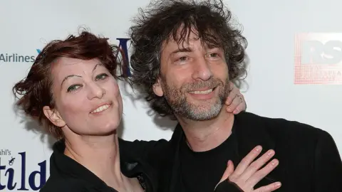 A mid shot: Palmer, a woman with tattooed eyebrows and dyed red hair, rests her hands on the chest and back of the neck of Gaiman, bearded with shaggy hair, as they pose, wearing black, at a media event