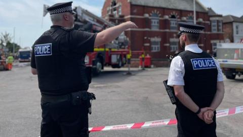 Two police officers, one in a black uniform and one wearing a black stab vest and a white shirt, stand in front of a fire engine on a street in Southport