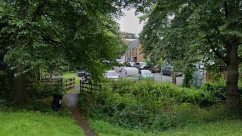 A footbridge over a stream overgrown with vegetation, showing houses in Banning Street in the background