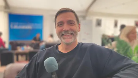 Patrick Grant - a man smiling whilst holding a microphone sat on a sofa.