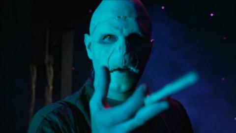A man wearing a bald cap holds a wand up to the camera. He has prosthetics on, so instead of a nose, there are two slits in his face