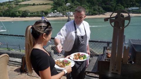 A chef serves food from a BBQ on a restaurant terrace by a river