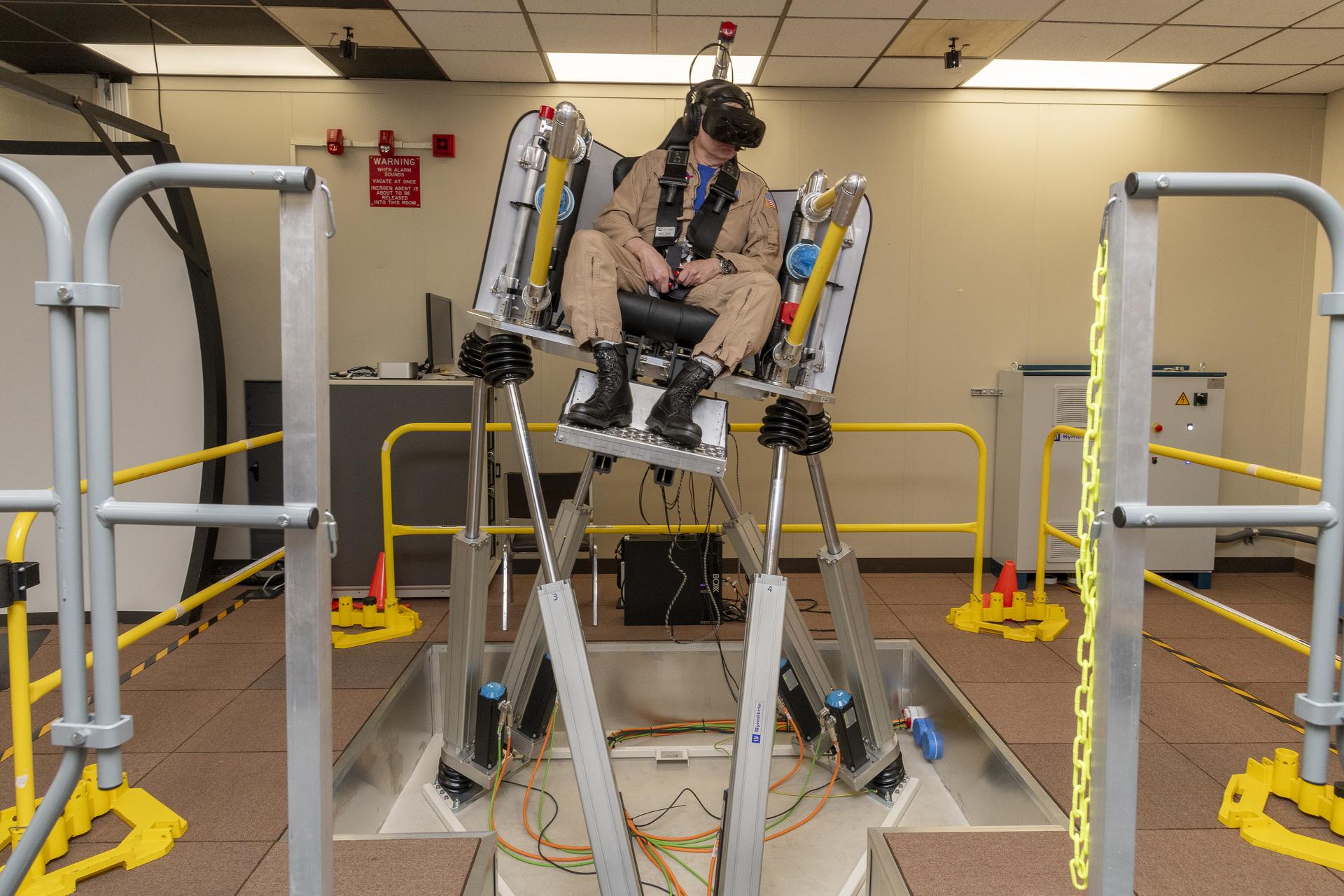 A man in a tan flight suit with black boots sits in a black seat on top of a metal platform below. He is strapped into the seat and wears a black headset and black, large goggles. He is tilted in the seat where the left side is angled down and the right side is angled up due to the motion of the simulator seat.